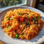 A close-up of a white plate filled with vibrant orange mexican rice recipe mixed with bright green peas and diced carrots.