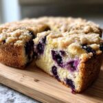 Close-up of a moist blueberry breakfast cake slice showing abundant blueberries and a thick crumb topping on a wooden board.