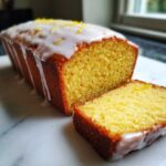 A close-up of a freshly baked lemon loaf with a thick white glaze and lemon zest, showing one slice cut ready to serve.