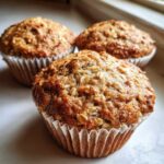 Three freshly baked, golden brown oatmeal muffins sitting on a white surface, with visible oat flakes on top.