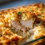 Close-up of a serving spoon lifting a piece of moist chicken from a creamy chicken and rice casserole with a golden, crispy breadcrumb topping.