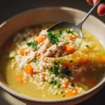 A spoonful of rich penicillin soup featuring shredded chicken, small pasta pearls, and diced carrots being lifted from a bowl.
