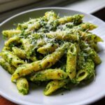 A close-up shot of penne pesto pasta coated in vibrant green sauce and topped with grated Parmesan cheese.