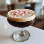 A rich peppermint espresso martini in a coupe glass, topped with a thick layer of foam and crushed candy canes.