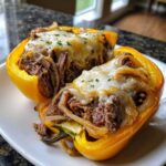 Two halves of a yellow bell pepper filled with shredded beef, onions, and topped with melted cheese, resembling philly cheesesteak stuffed peppers.