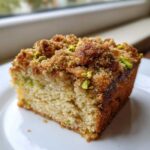 A moist slice of pistachio coffee cake featuring a thick, sugary brown crumb topping sprinkled with bright green chopped pistachios.