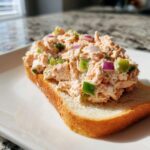 A generous scoop of creamy salmon salad with visible chunks of red onion and green pepper served open-faced on toasted white bread.