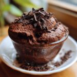 A close-up of rich, dark chocolate mousse topped with chocolate shavings, served in a small glass cup.