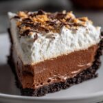 Close-up of a decadent slice of mud pie showing a dark crust, thick chocolate layer, and fluffy white topping garnished with shavings.