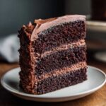 A tall slice of moist, three-layer chocolate cake with thick chocolate frosting, served on a white plate.