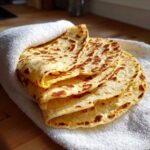 A stack of freshly cooked, soft corn tortillas wrapped partially in a white towel on a wooden surface.