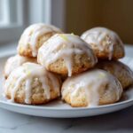 A close-up of soft, dome-shaped ricotta cookies piled on a white plate, generously drizzled with white lemon glaze and topped with zest.