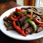 Close-up of sizzling steak fajitas featuring grilled strips of steak, red bell peppers, green bell peppers, and onions on a white plate.