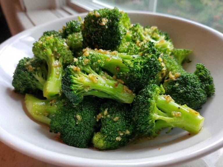 Close-up of bright green steamed broccoli florets tossed with minced garlic and seasoning in a white bowl.