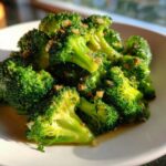 A close-up of bright green steamed broccoli florets tossed in garlic and chili flakes, served on a white plate.