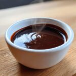 Close-up of rich, dark chocolate fondue steaming slightly in a small white bowl on a wooden surface.
