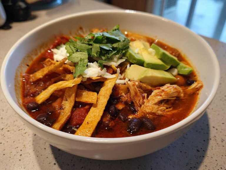 A close-up of a bowl filled with rich, red broth chicken tortilla soup recipe, topped with avocado, cheese, cilantro, and crispy tortilla strips.