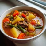 Close-up of a white bowl filled with rich vegetable beef soup, featuring chunks of potato, carrot, green beans, and shredded beef.