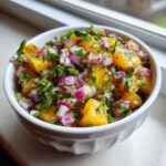 Close-up of a white bowl filled with fresh peach salsa, featuring diced yellow peaches, red onion, and cilantro.