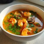 Close-up of a white bowl filled with bright orange tom yum soup, featuring large shrimp and sliced mushrooms, garnished with fresh cilantro.