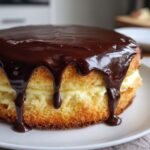 A beautiful, tall slice of boston cream pie featuring golden sponge cake, thick vanilla custard filling, and rich chocolate ganache dripping down the sides.