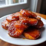 Close-up of shiny, caramelized candied yams stacked on a white plate near a window.