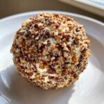A perfectly formed cheese ball coated entirely in chopped pecans, sitting on a white plate.