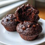 A stack of three rich, dark chocolate muffins topped with chocolate chips, one is broken open showing the moist interior.