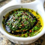 Close-up of a white bowl containing olive oil bread dip with herbs, chili flakes, and balsamic vinegar swirl.