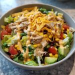 A close-up view of a hearty southwest salad topped with shredded chicken, black beans, corn, avocado, and creamy dressing.