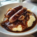 Two browned sausages served over creamy mashed potatoes, smothered in rich onion gravy, a classic bangers and mash dish.