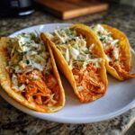 Three crispy shell buffalo chicken tacos topped with creamy coleslaw on a white plate.