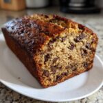 A close-up of a freshly baked loaf of protein banana bread studded with chocolate chips, resting on a white plate.