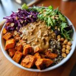 Close-up of a vibrant buddha bowl featuring roasted sweet potatoes, greens, red cabbage, chickpeas, quinoa, and peanut sauce.