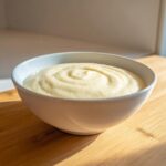 A close-up of thick, creamy cauliflower soup swirled in a white bowl, sitting on a wooden surface.