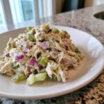 A serving of creamy, shredded chicken protein salad mixed with diced green celery and purple red onion on a white plate.