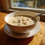 A creamy bowl of chicken and rice soup sits on a wooden table near a window, perfect for a comforting rice soup.