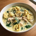 Close-up of creamy chicken tortellini soup with shredded chicken, spinach, and spices in a white bowl.