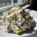A mound of creamy cottage cheese chicken salad featuring shredded chicken, bright green celery, and purple onion chunks on a white plate.