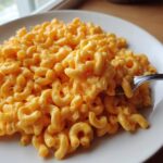 A close-up of creamy, bright orange cottage cheese mac and cheese served on a white plate with a fork lifting some pasta.