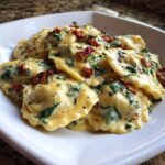 A close-up of a white bowl filled with creamy Tuscan ravioli mixed with spinach and topped with sun-dried tomatoes.