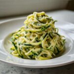 A mound of creamy zucchini noodles coated in a light sauce, served on a white plate.