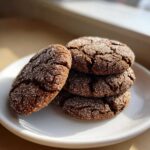 A stack of four rich, dark molasses cookies covered in sparkling sugar crystals, sitting on a white plate.