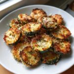 A plate piled high with golden brown, crispy roasted zucchini slices topped with Parmesan cheese and herbs.