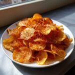 A mound of golden, crispy homemade potato chips seasoned with spices, piled high on a white plate.