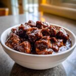 Close-up of tender crockpot beef tips coated in a thick, dark, glossy sauce served in a white bowl.