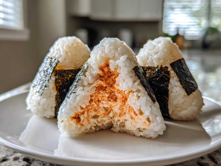 Three triangular tuna rice balls (onigiri) on a white plate, one is bitten to show the spicy orange tuna filling.