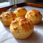 Four golden-brown, fluffy carnivore bread muffins cooling on parchment paper near a window.