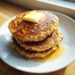 A delicious stack of three fluffy oatmeal pancakes topped with melting butter and syrup.