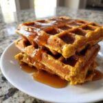 A close-up of a stack of fluffy pumpkin waffles generously drizzled with amber syrup.